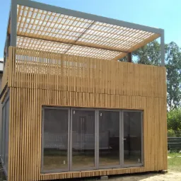 Nowoczesny, modułowy dom z drewnianą elewacją i panoramicznymi oknami, zwieńczony ażurową pergolą na dachu, widoczny w letnim słońcu.