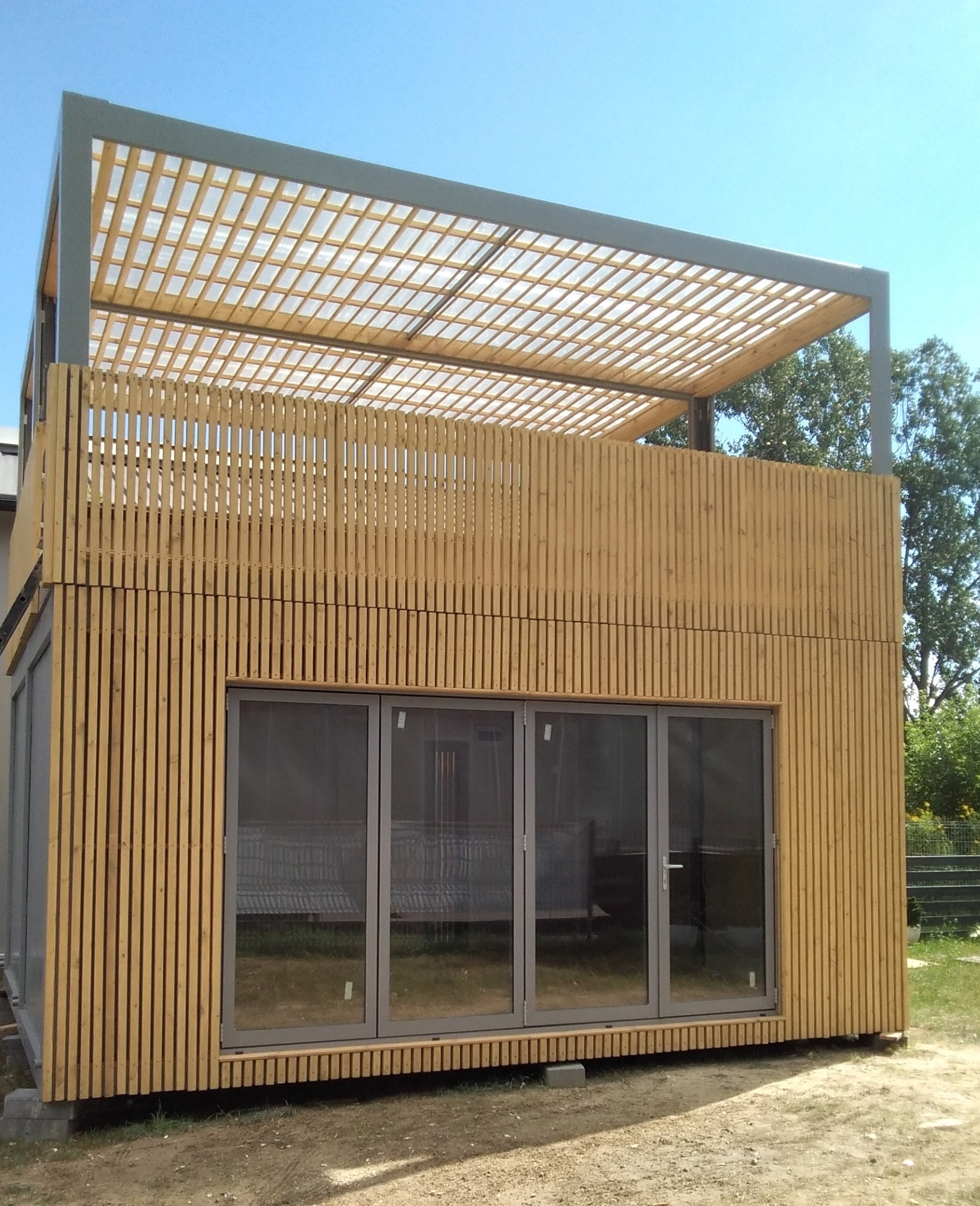 Nowoczesny, modułowy dom z drewnianą elewacją i panoramicznymi oknami, zwieńczony ażurową pergolą na dachu, widoczny w letnim słońcu.