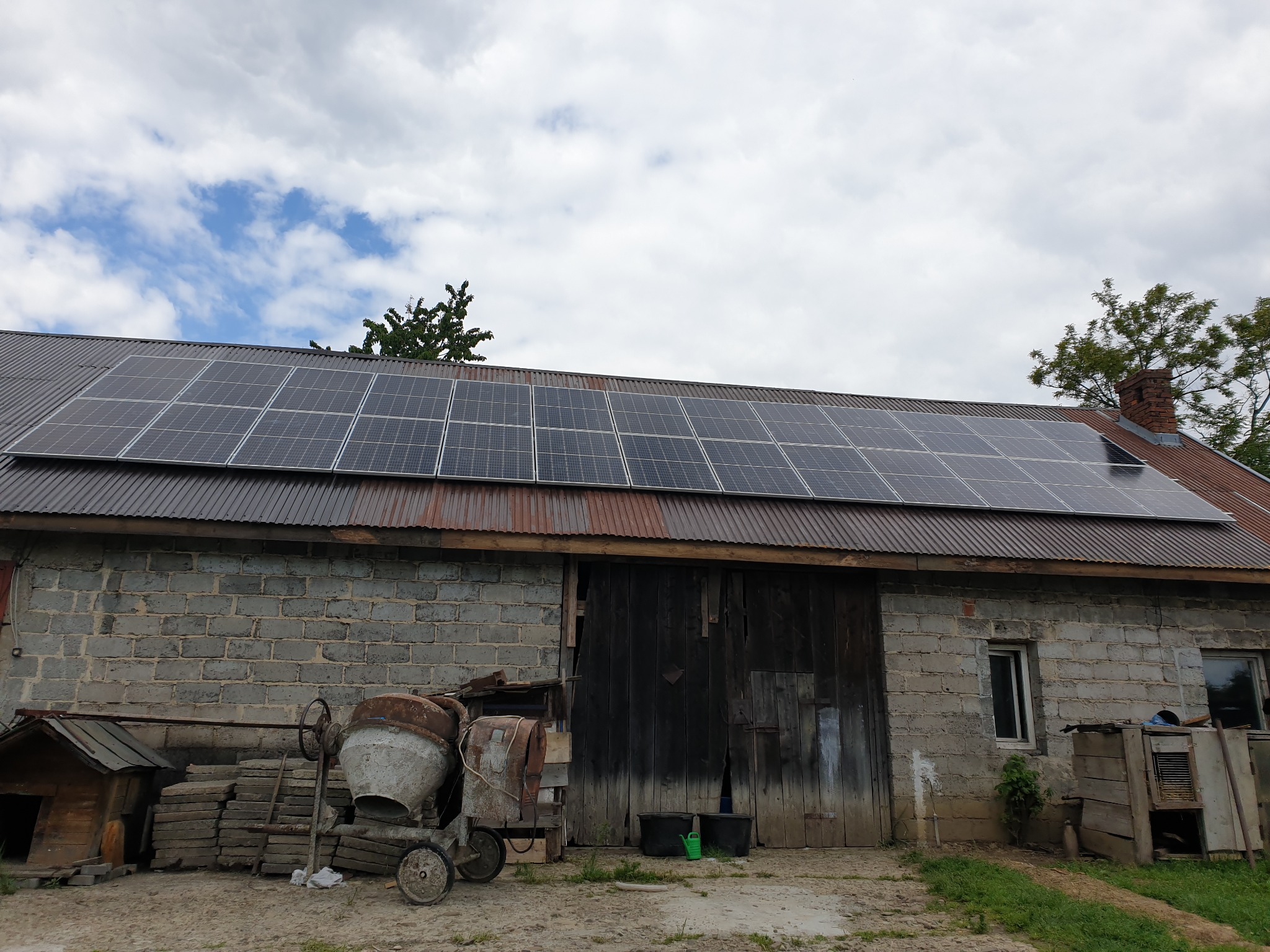 Panele słoneczne na dachu starego, murowanego budynku gospodarczego z betonową mieszarką i stertą płyt chodnikowych w pobliżu.