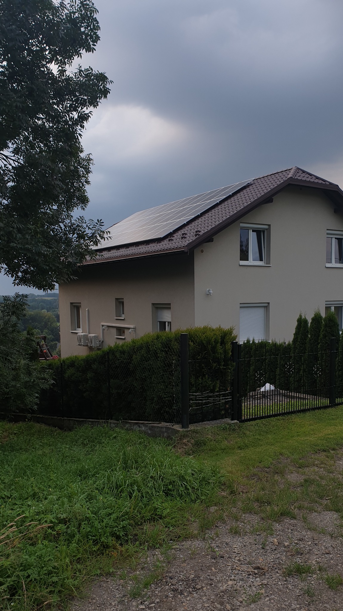 Dwupiętrowy dom z beżową elewacją i brązowym dachem, na którym zamontowane są panele słoneczne. Widok z poziomu gruntu, z fragmentem drzewa po lewej stronie i zielenią w dolnej części kadru. Za...