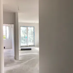 Wnętrze domu w stanie deweloperskim: surowe ściany, wylewka na podłodze, okna zabezpieczone folią, widoczne instalacje elektryczne.