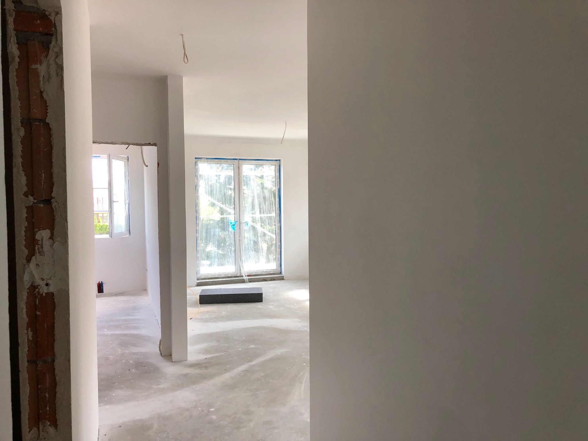 Wnętrze domu w stanie deweloperskim: surowe ściany, wylewka na podłodze, okna zabezpieczone folią, widoczne instalacje elektryczne.