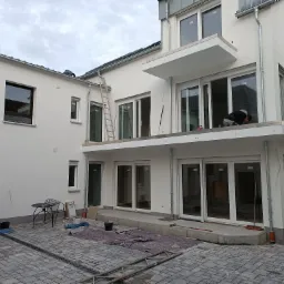 Biały, dwupiętrowy dom w trakcie prac wykończeniowych elewacji, z widocznym pracownikiem na balkonie, drabina oparta o ścianę i rozłożone narzędzia na brukowanym podwórku.