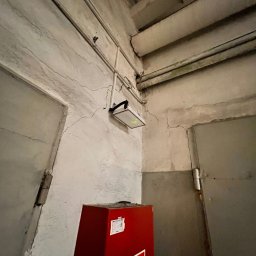 Igor Jasiński Electric Solutions - Awaryjne oświetlenie LED zamontowane na ścianie w pobliżu skrzynki hydrantowej wewnątrz budynku, widoczne pęknięcia na ścianie.