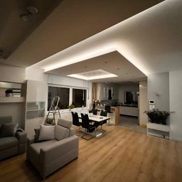 Igor Jasiński Electric Solutions - Nowoczesny salon z oświetleniem LED w suficie podwieszanym, widoczna drabina oparta o okno, sugerująca prace wykończeniowe lub instalacyjne.