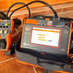 Igor Jasiński Electric Solutions - Miernik elektryczny Sonel MPI-540 w torbie, z podłączonym adapterem EVSE-01, wyświetlający wyniki pomiarów na ekranie LCD, na tle ceramicznej podłogi i drewnianego panelu.