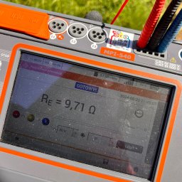 Igor Jasiński Electric Solutions - Miernik MPI-540 z podłączonymi przewodami pomiarowymi, ekran wyświetla wynik rezystancji uziemienia 9,71 omów, w tle zielona trawa.