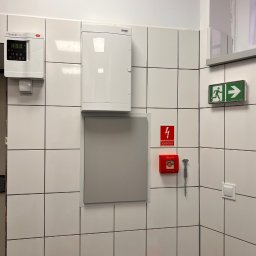 Igor Jasiński Electric Solutions - Biała ściana w kafelkach z zamontowaną skrzynką elektryczną, panelem sterowania, przyciskiem alarmowym i znakiem ewakuacyjnym.