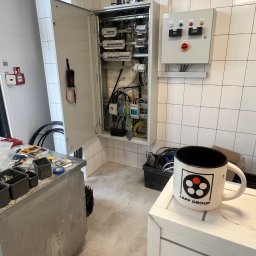 Igor Jasiński Electric Solutions - Otwarta rozdzielnica elektryczna z widocznymi przewodami i bezpiecznikami, obok panel sterowania na kafelkowej ścianie. Na pierwszym planie narzędzia i kubek LAPP GROUP.