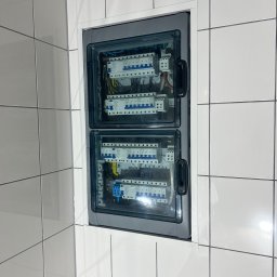 Igor Jasiński Electric Solutions - Wbudowana dwusekcyjna rozdzielnica elektryczna Legrand z niebieskimi i białymi wyłącznikami, osadzona w ścianie z białych, błyszczących płytek z ciemną fugą.