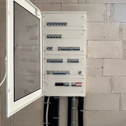 Igor Jasiński Electric Solutions - Biała rozdzielnica elektryczna z otwartymi drzwiczkami, ukazująca szereg niebieskich wyłączników nadprądowych, czerwony różnicowoprądowy i cyfrowy wyświetlacz, zamontowana na ścianie z jasnych...