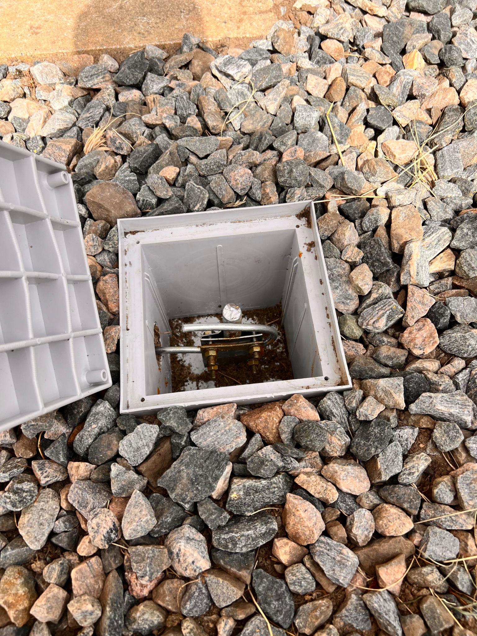 Otwarta, kwadratowa studzienka rewizyjna z metalową konstrukcją wewnątrz, otoczona dekoracyjnymi kamieniami.