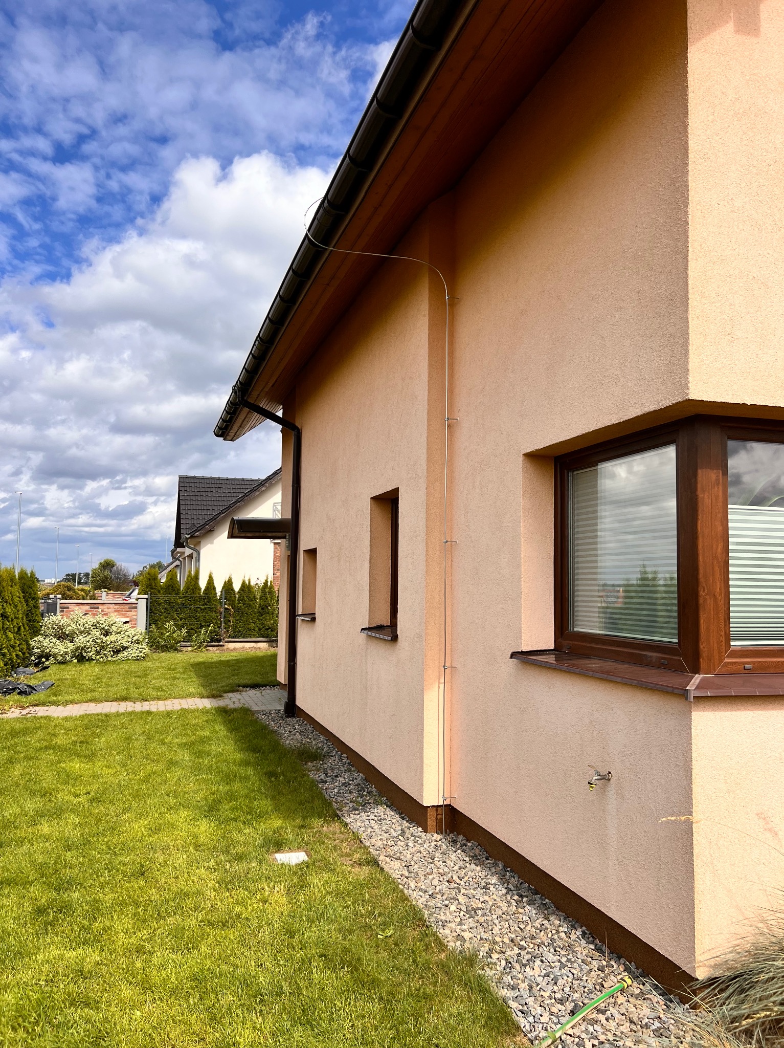 Elewacja domu z instalacją odgromową poprowadzoną po ścianie, widoczna rura spustowa i okno z żaluzjami.