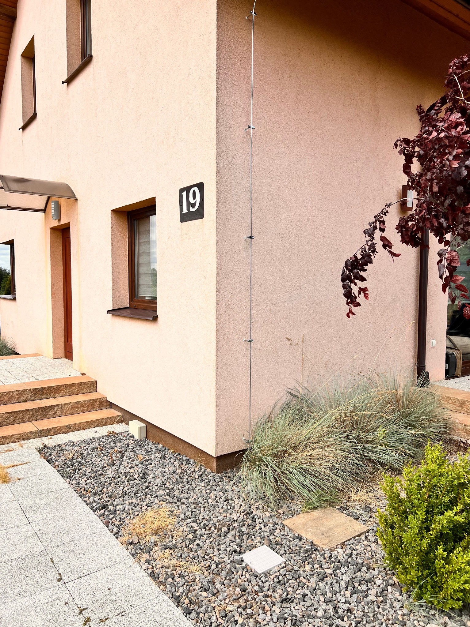 Elewacja domu z numerem 19, widoczna pionowa instalacja z metalowymi uchwytami, prawdopodobnie odprowadzająca ładunki elektryczne, obok wejścia z kamiennymi schodami i ozdobne krzewy.