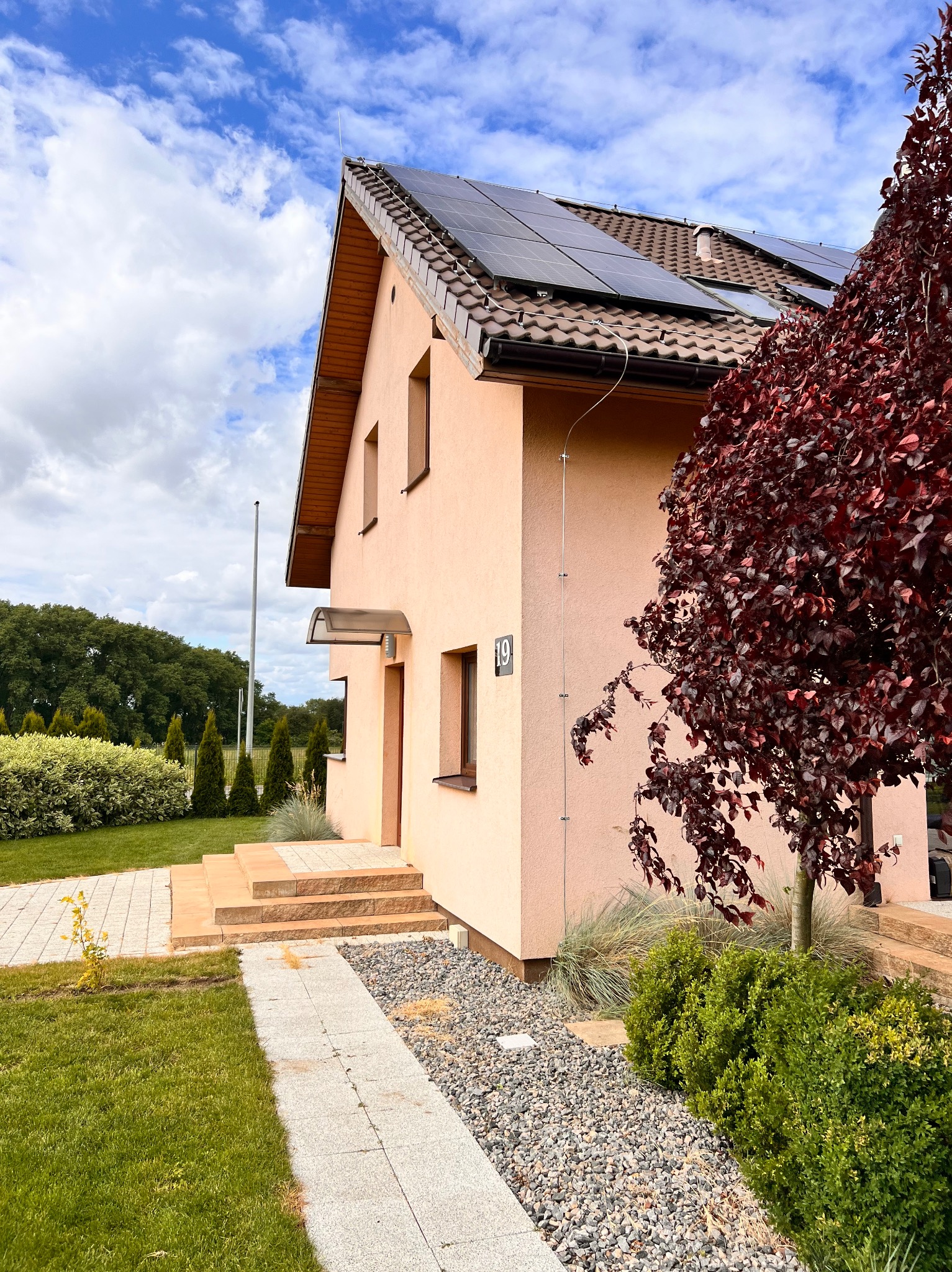 Dom jednorodzinny z panelami słonecznymi na dachu i widoczną instalacją odgromową w postaci pionowego przewodu na elewacji, obok drzewo o purpurowych liściach i chodnik z kostki brukowej.