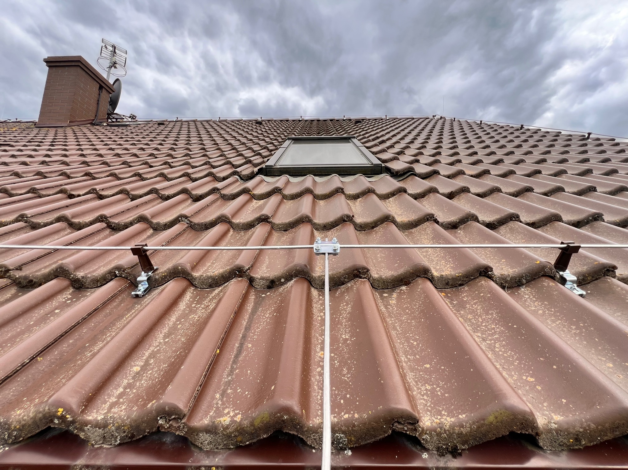 Ujęcie z dołu na brązowy dach z dachówką karpiówką, na którym zamontowano system odgromowy z metalowymi uchwytami i przewodami, widoczne okno dachowe, komin z anteną i pochmurne niebo.