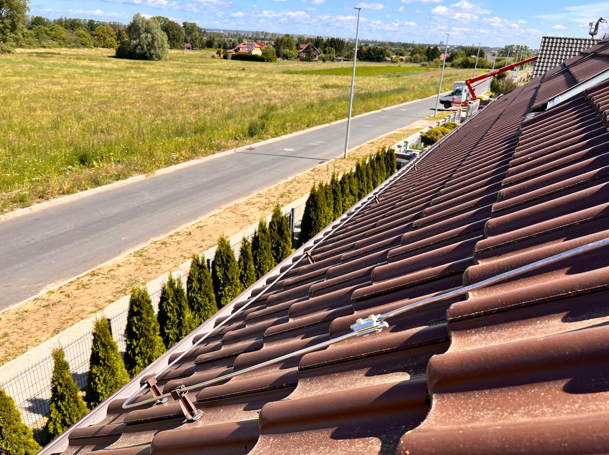 Brązowy dach z zamontowanym systemem ochrony odgromowej, widok z góry na ulicę, pole i oddalony podnośnik koszowy.