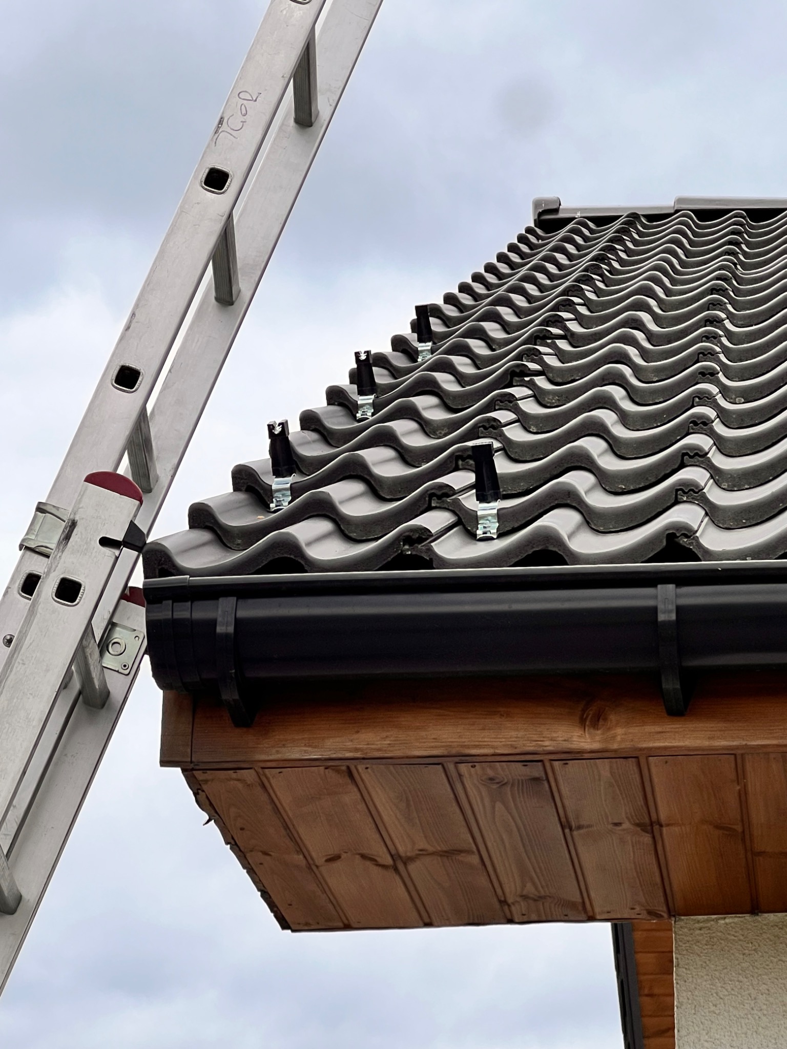 Szare dachówki z zamontowanymi uchwytami odgromowymi, widoczne z drabiny przystawionej do dachu i rynny w kolorze czarnym.