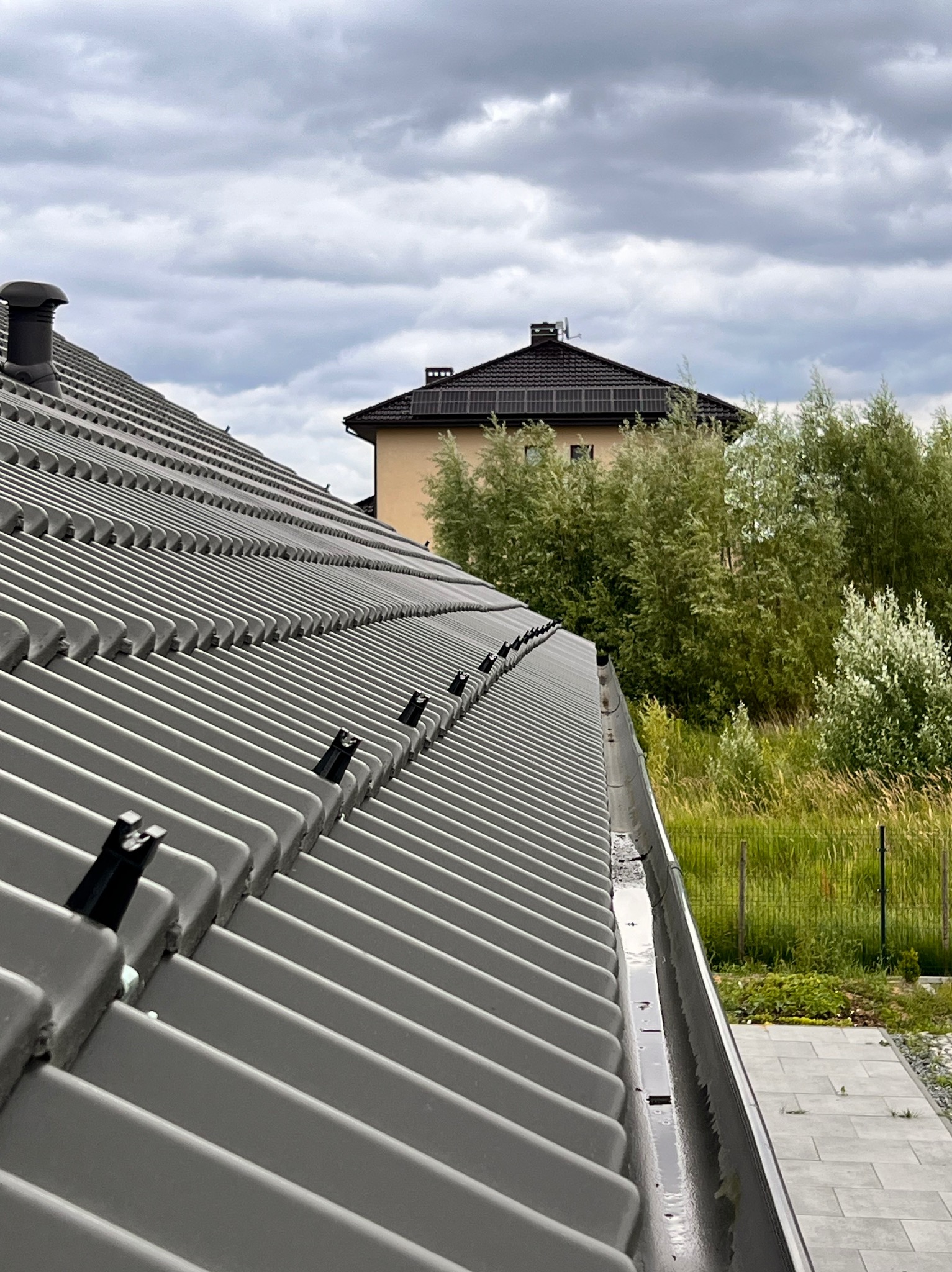 Szary dach z blachy trapezowej ze wspornikami instalacji odgromowej w Szczecinie, widok z góry na tle zieleni i innego budynku pod pochmurnym niebem.