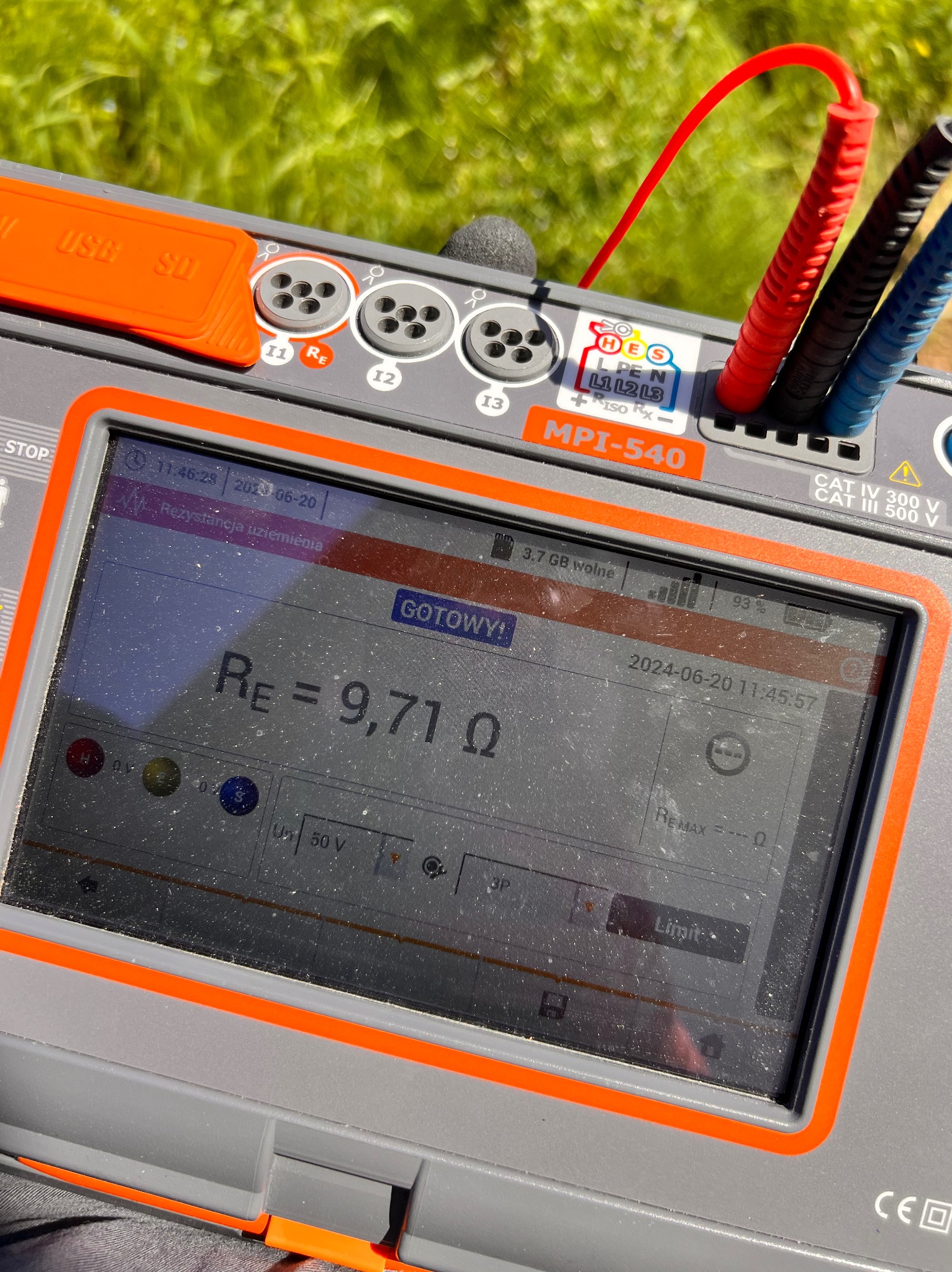 Miernik MPI-540 z podłączonymi przewodami pomiarowymi, ekran wyświetla wynik rezystancji uziemienia 9,71 omów, w tle zielona trawa.