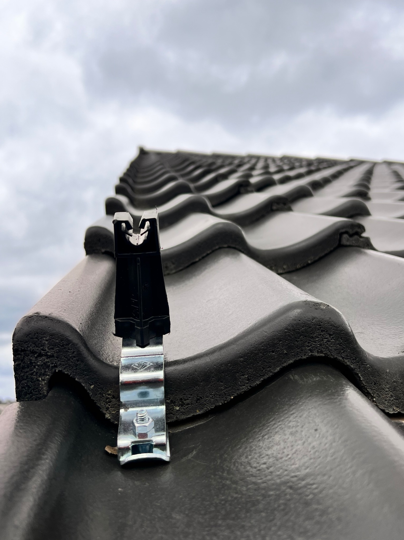 Metalowy uchwyt mocujący na ciemnej dachówce ceramicznej, zamocowany śrubą, widok z dołu na pochmurne niebo.