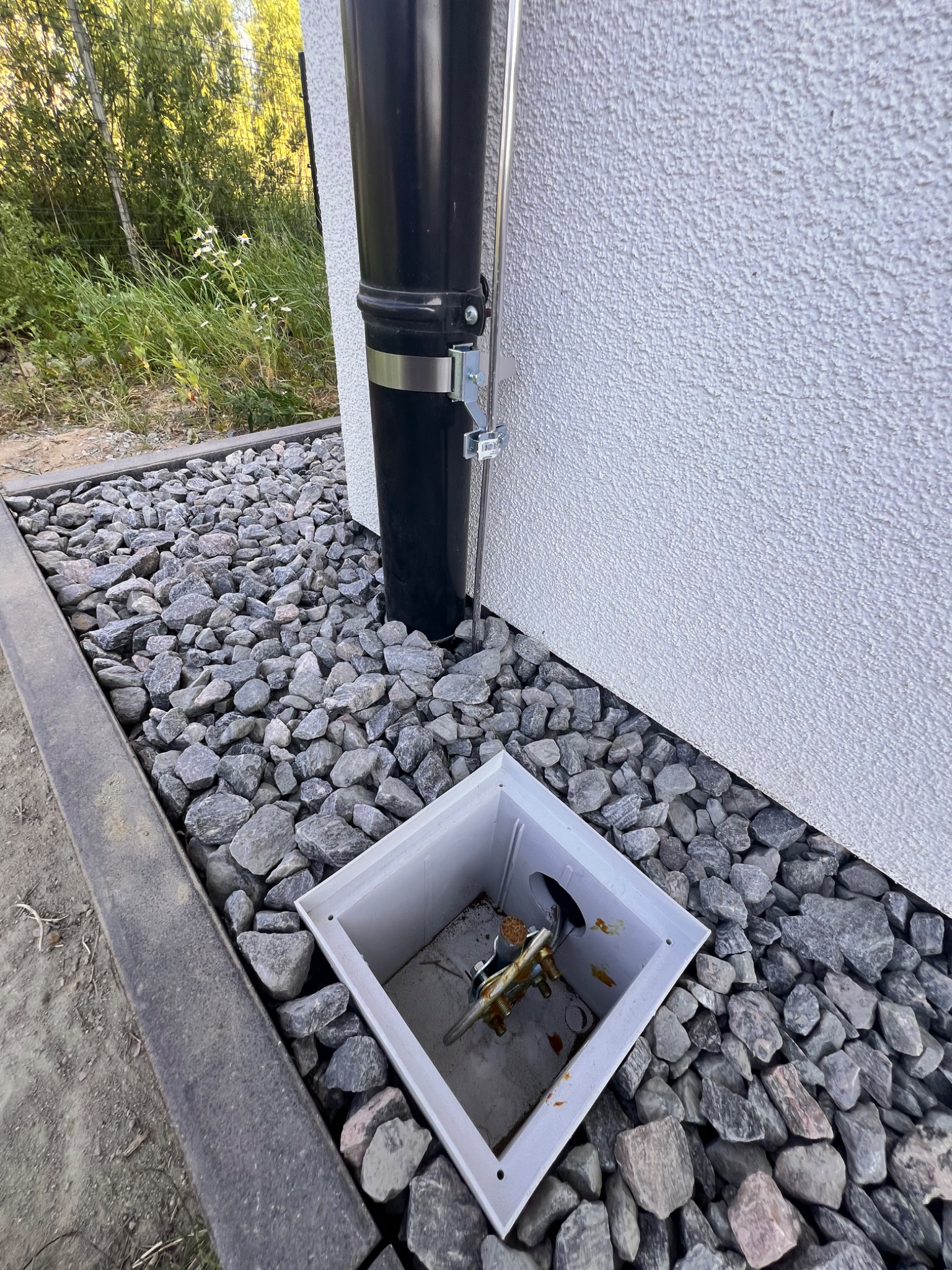 Uziemienie instalacji odgromowej ukryte w białej skrzynce rewizyjnej, widoczny przewód odprowadzający przy rynnie, otoczenie wysypane szarymi kamieniami.