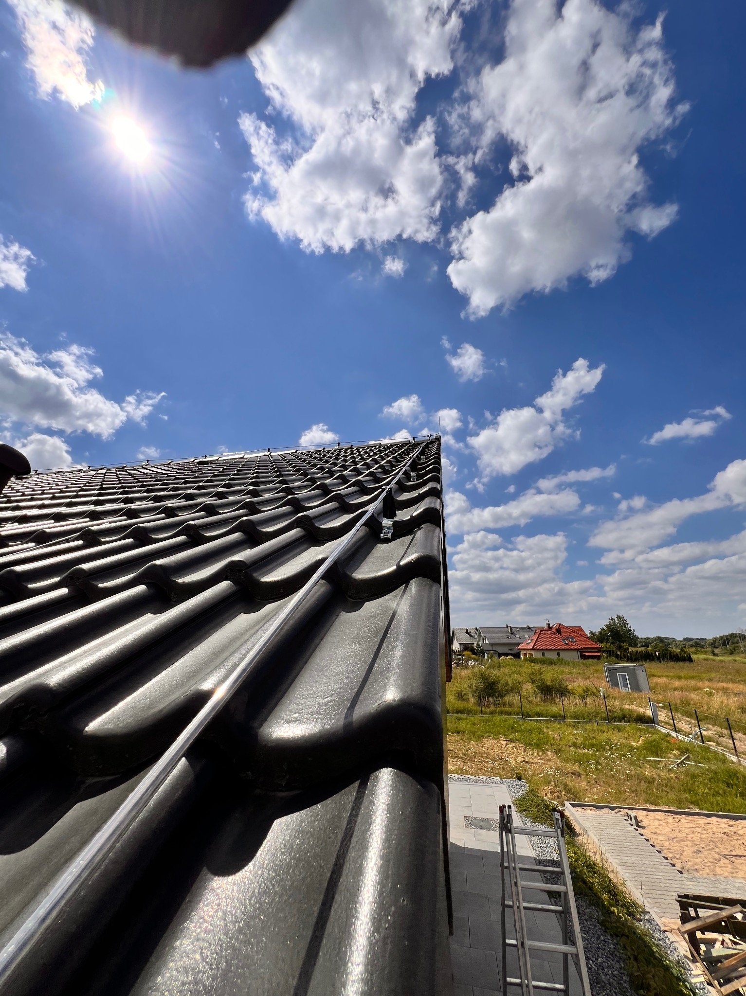 Czarny dach z dachówki ceramicznej z zamontowaną instalacją odgromową, widok z dołu na tle błękitnego nieba z chmurami i krajobrazu z domami.
