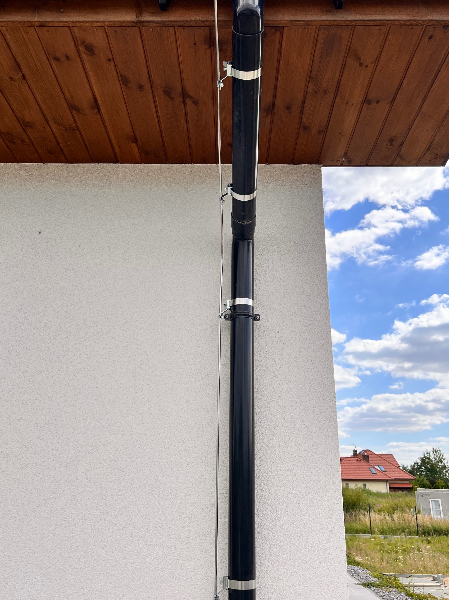 Czarna rura spustowa z metalowymi obejmami przymocowana do jasnej ściany budynku, obok widoczny przewód odgromowy, w tle błękitne niebo z chmurami i fragment dachu sąsiedniego domu.