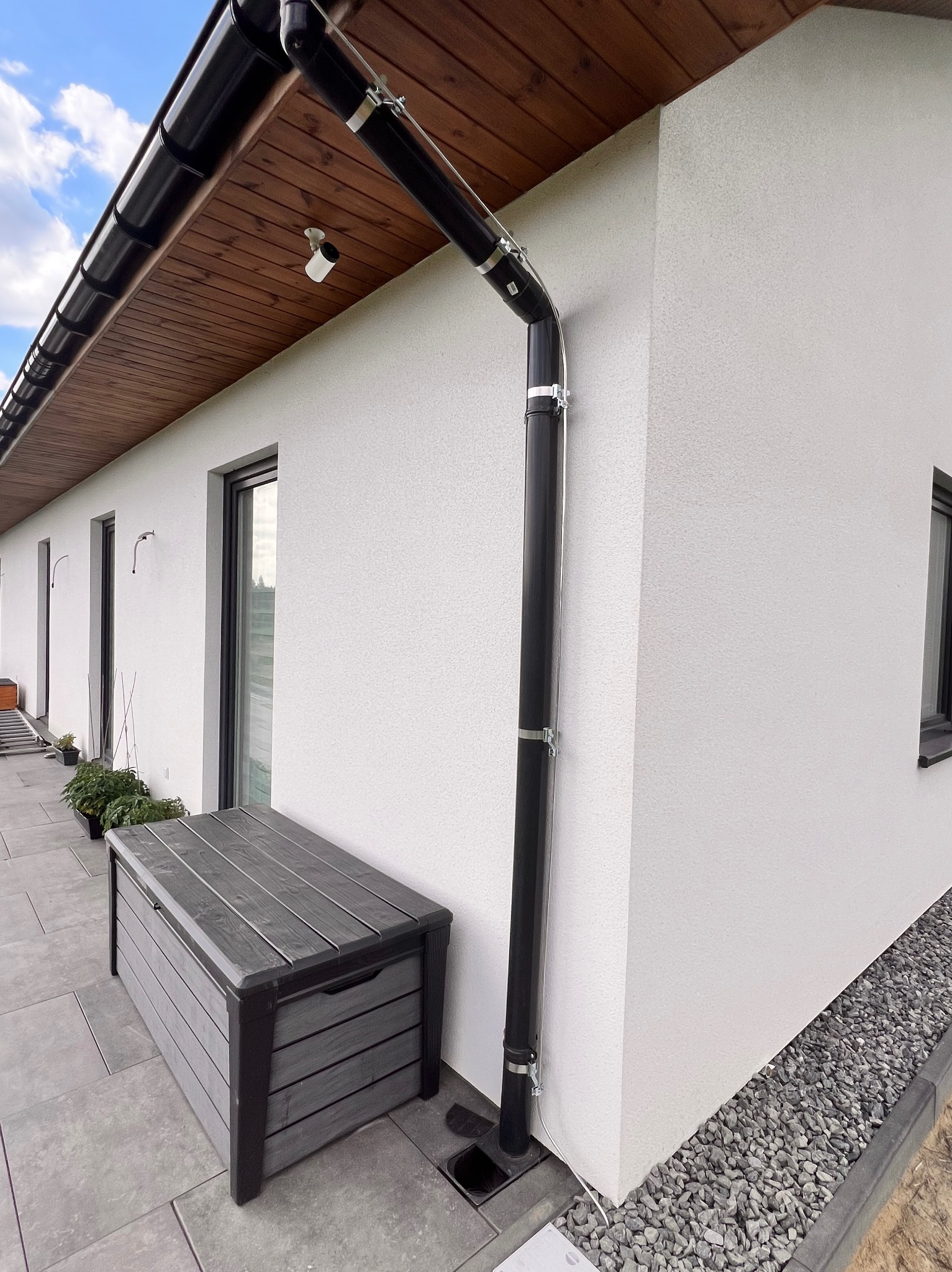 Czarna rynna pionowa przymocowana do białej ściany domu, z widocznym czarnym odpływem, skrzynią ogrodową i szarą podsypką z kamienia.