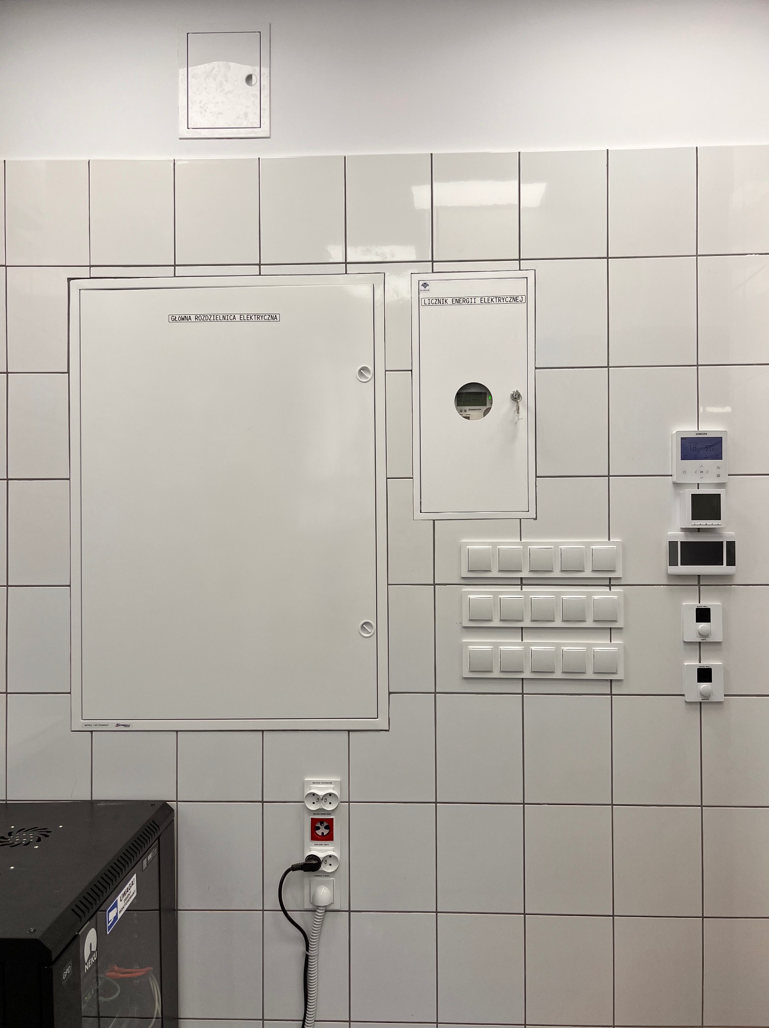 Biała ściana w płytkach z zamontowaną główną rozdzielnicą elektryczną, licznikiem energii, włącznikami światła i gniazdkami. Widoczny fragment szafy serwerowej.