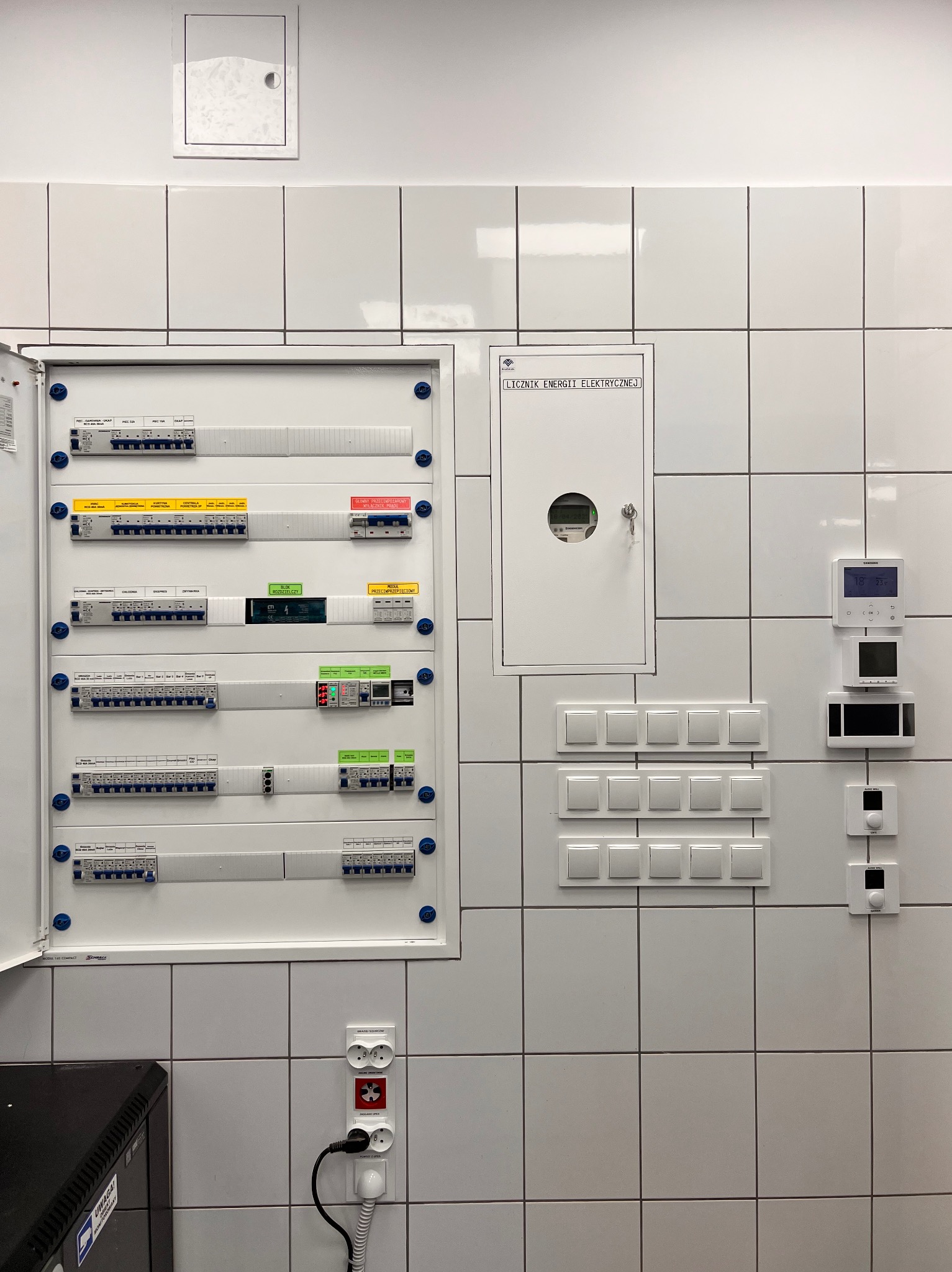 Otwarta skrzynka elektryczna z bezpiecznikami na kafelkowanej ścianie, obok licznika energii i włączników światła. Widoczne gniazdka i podłączony kabel. Białe tło.