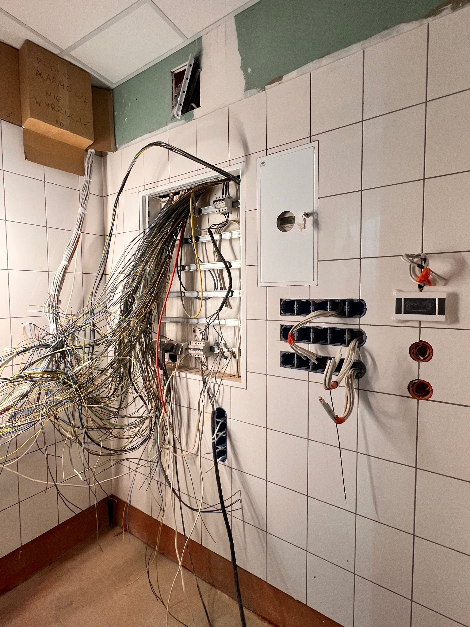 Ściana w kafelkach z rozciągniętymi kablami elektrycznymi w trakcie instalacji, widoczne puszki i skrzynka bezpiecznikowa. Częściowo wykończone wnętrze.
