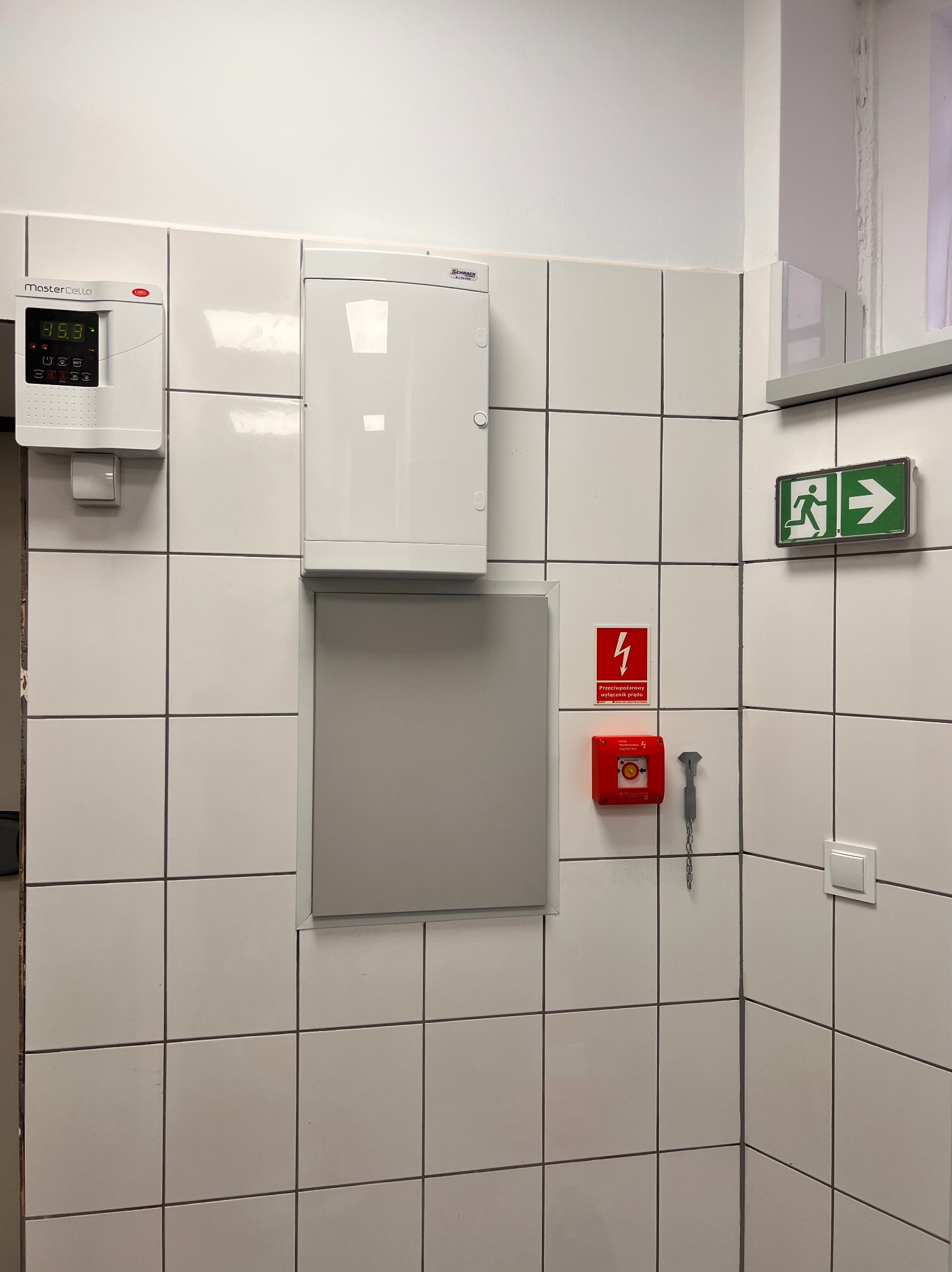Biała ściana w kafelkach z zamontowaną skrzynką elektryczną, panelem sterowania, przyciskiem alarmowym i znakiem ewakuacyjnym.