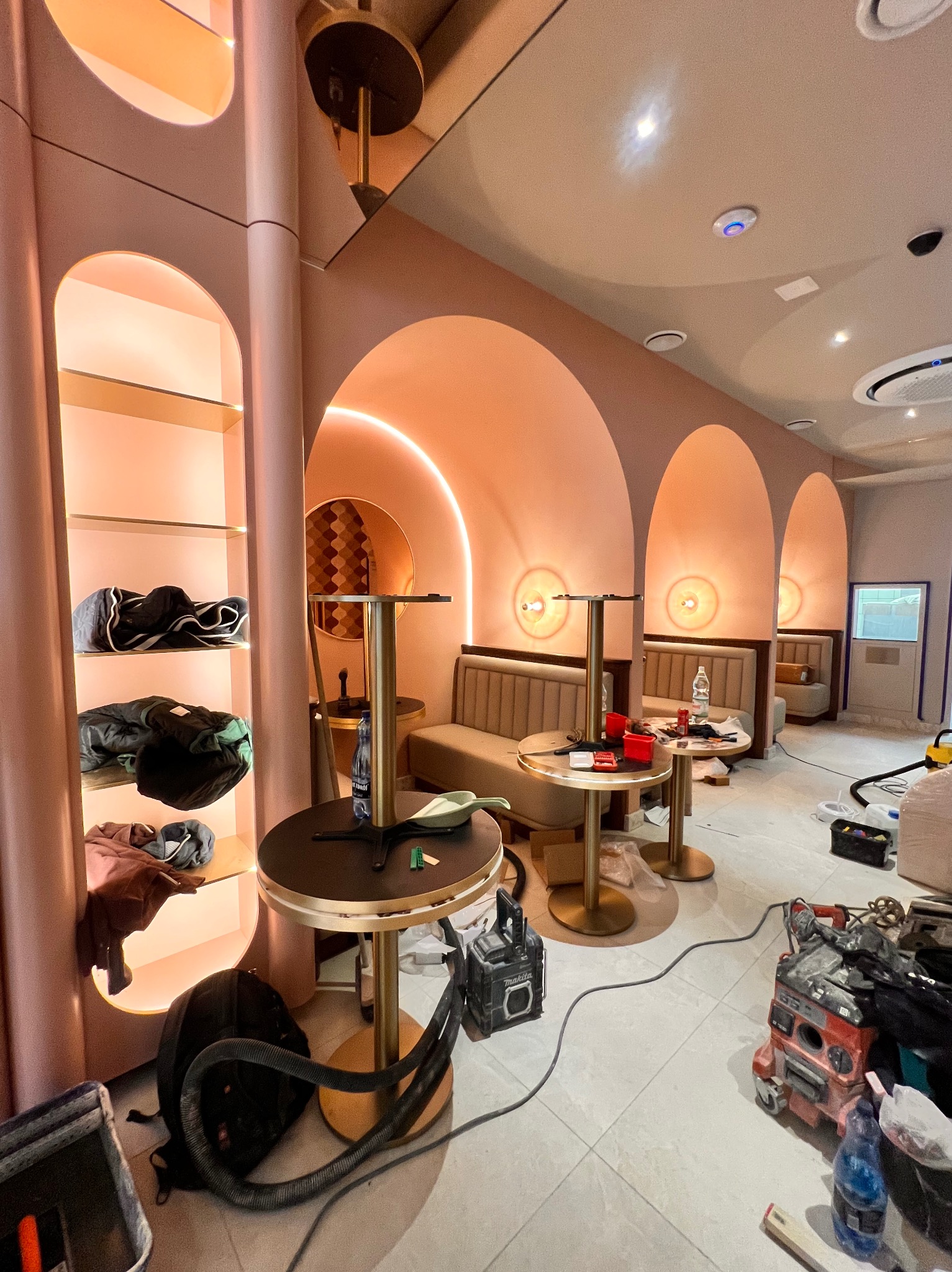 Wnętrze lokalu w trakcie prac wykończeniowych: widoczne stoliki barowe, oświetlenie w łukach i narzędzia budowlane. Elementy w odcieniach różu i złota.