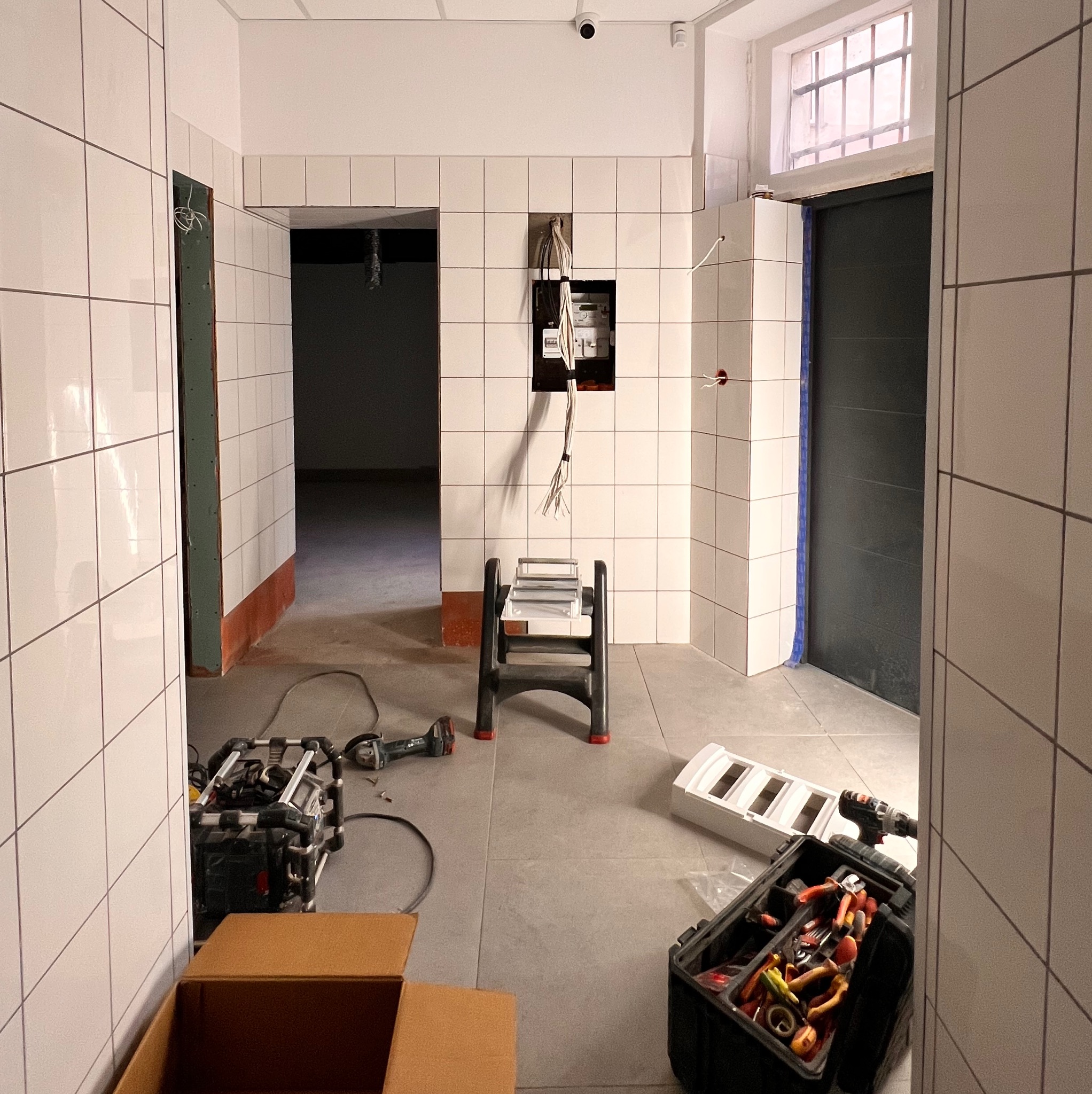 Wnętrze w trakcie remontu: rozdzielnica elektryczna z wystającymi kablami, drabina, narzędzia, płytki na ścianach. Widoczny etap prac instalacyjnych.