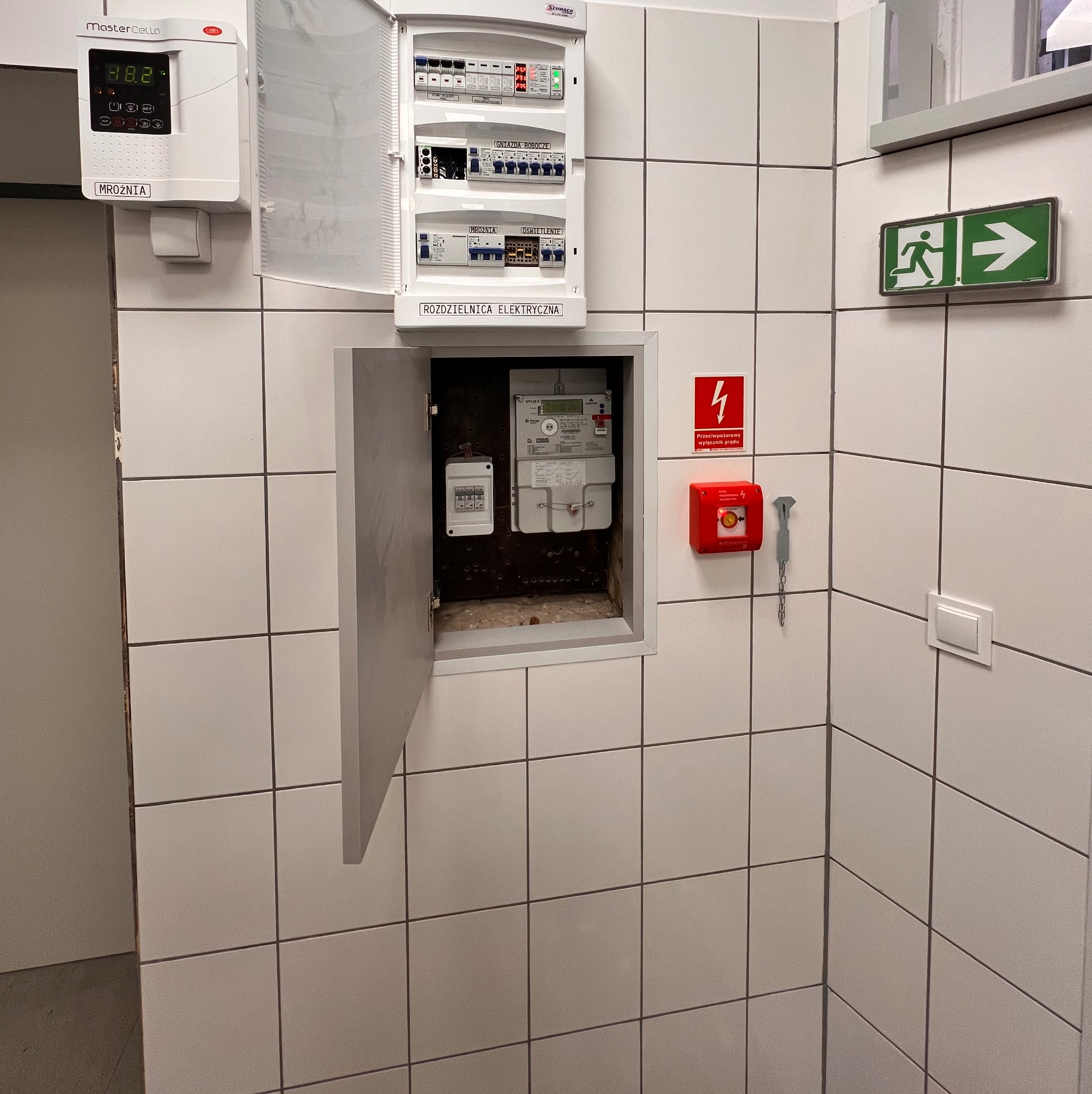Otwarta szafka z licznikiem elektrycznym, rozdzielnica elektryczna z bezpiecznikami, wyłącznik prądu i tablica informacyjna na ścianie z płytek w pomieszczeniu.