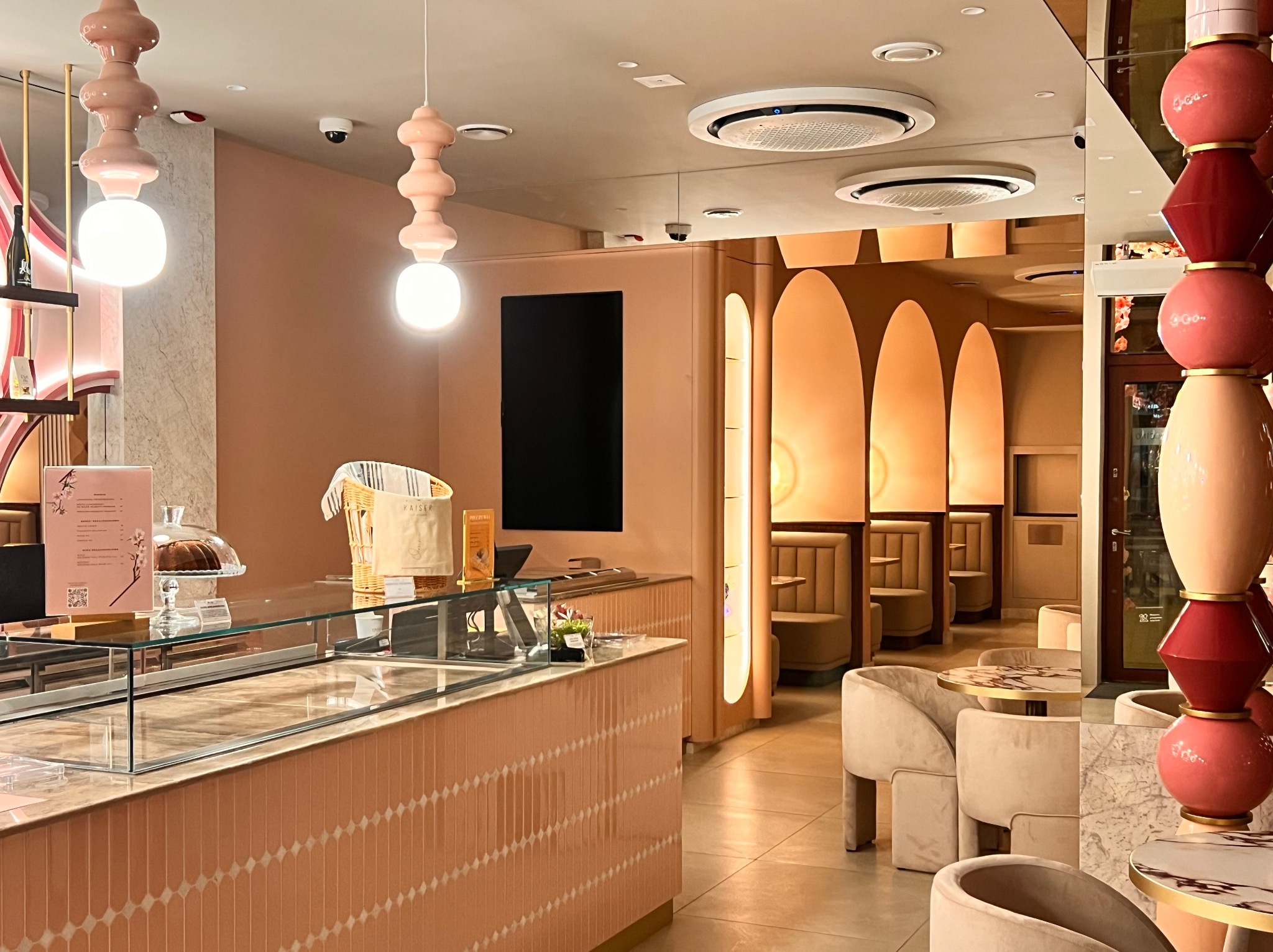 Nowoczesne oświetlenie w pastelowym wnętrzu kawiarni: wiszące lampy kuliste, okrągłe klimatyzatory sufitowe i kamery monitoringu. Widok na stoliki i kabiny w tle.