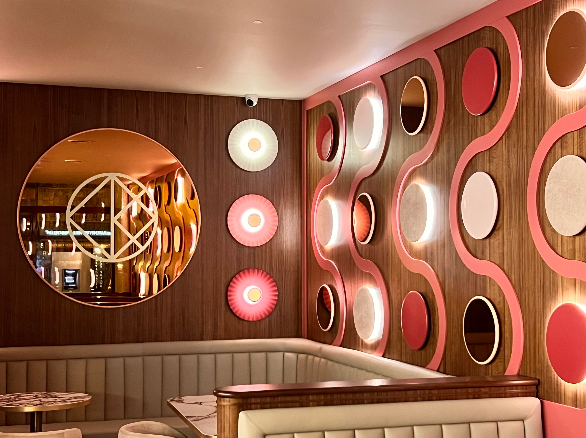 Stylowe oświetlenie dekoracyjne: okrągłe lampy LED w odcieniach różu i bieli na drewnianej ścianie, odbicie w lustrze i kamera przemysłowa w rogu.