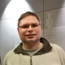 Portret mężczyzny w okularach i szarej bluzie z zamkiem błyskawicznym, na tle ściany z subtelnym wzorem postaci.