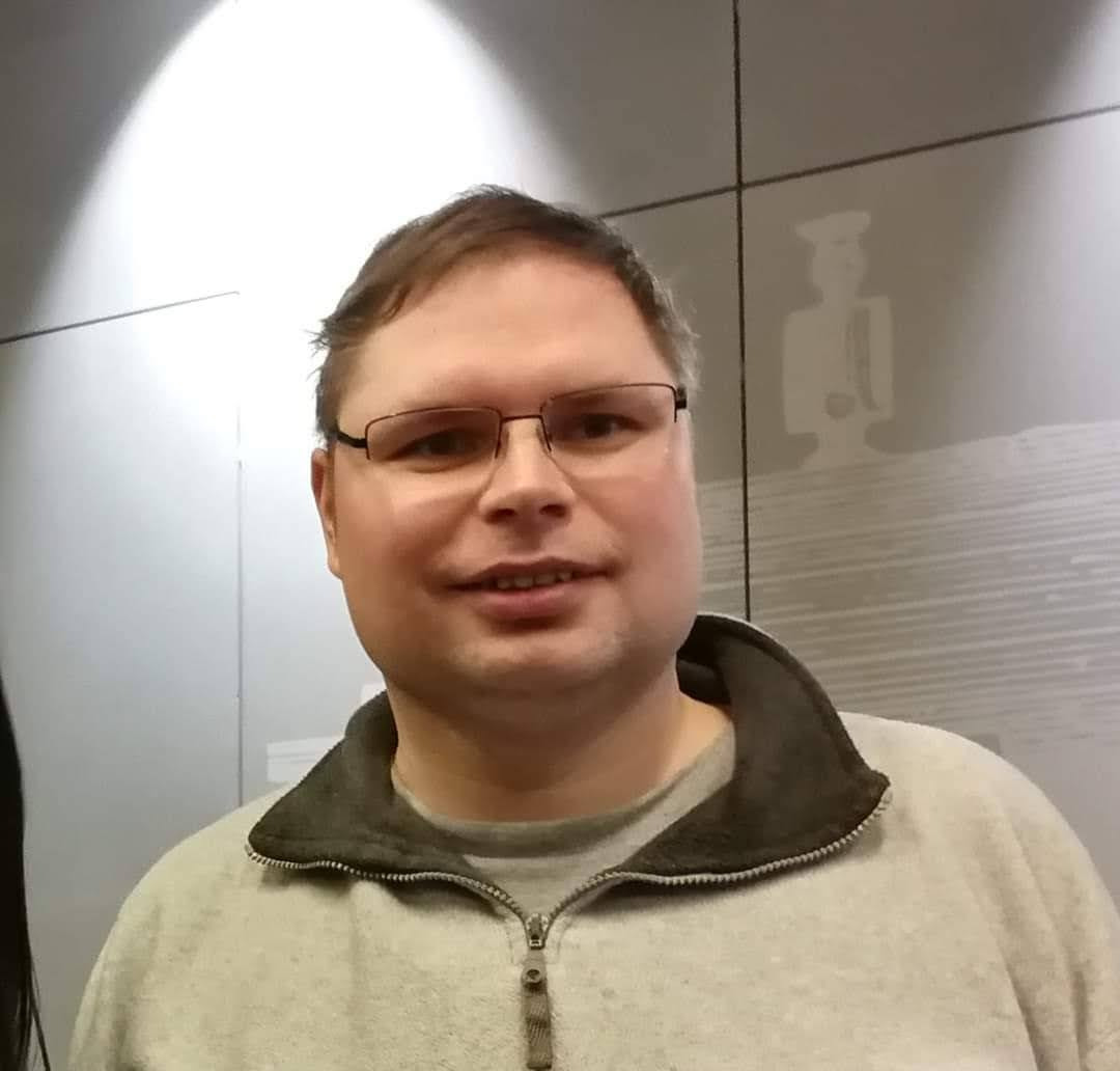 Portret mężczyzny w okularach i szarej bluzie z zamkiem błyskawicznym, na tle ściany z subtelnym wzorem postaci.