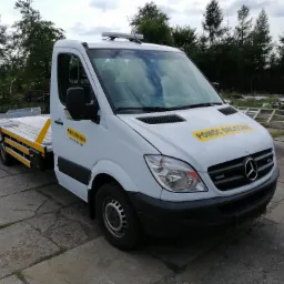 Biała laweta Mercedes-Benz z żółtymi napisami 'Pomoc Drogowa' zaparkowana na betonowym placu, w tle drzewa i stos opon.