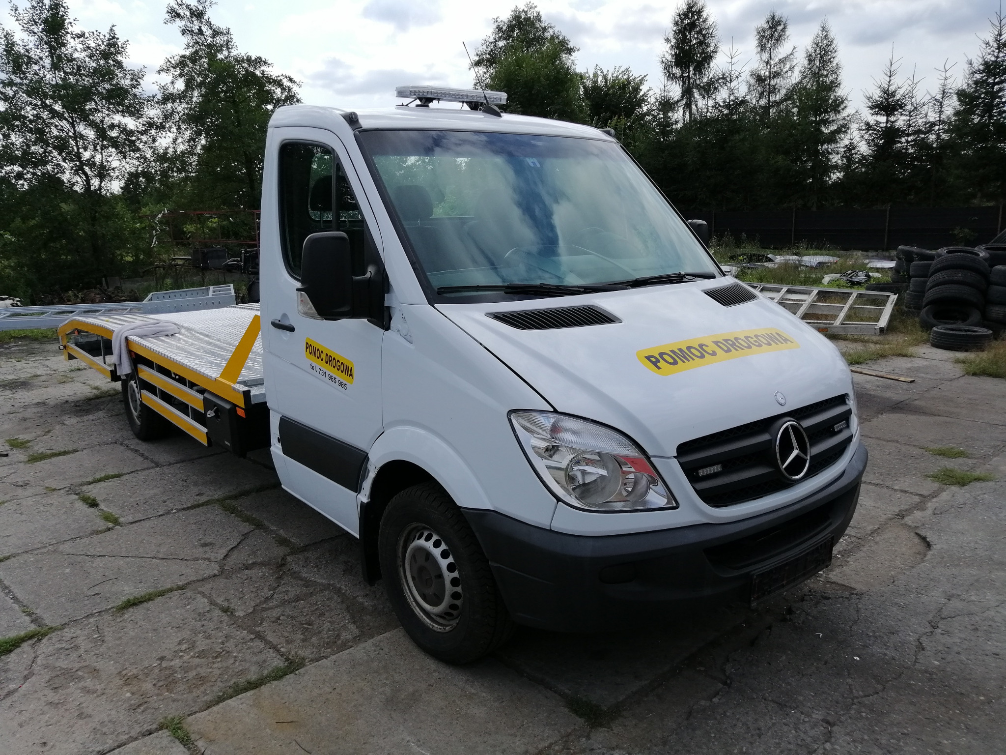 Biała laweta Mercedes-Benz z żółtymi napisami 'Pomoc Drogowa' zaparkowana na betonowym placu, w tle drzewa i stos opon.