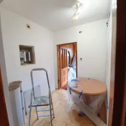 Przygotowane do odświeżenia wnętrze pokoju: ściany po nałożeniu gładzi, zabezpieczony folią stół, drabina malarska i rolki z materiałem ochronnym na parkiecie.