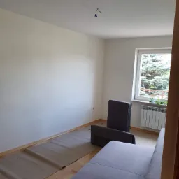 Pokój przygotowany do prac wykończeniowych: podłoga zabezpieczona kartonem, ściany zagruntowane, widoczne kable od lampy sufitowej.