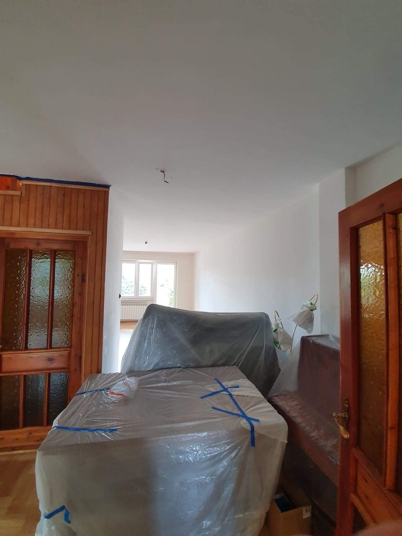 Przygotowane do odświeżenia wnętrze w Radomiu: meble zabezpieczone folią malarską, świeżo pomalowane ściany i sufit, drewniana boazeria z niebieską taśmą malarską.