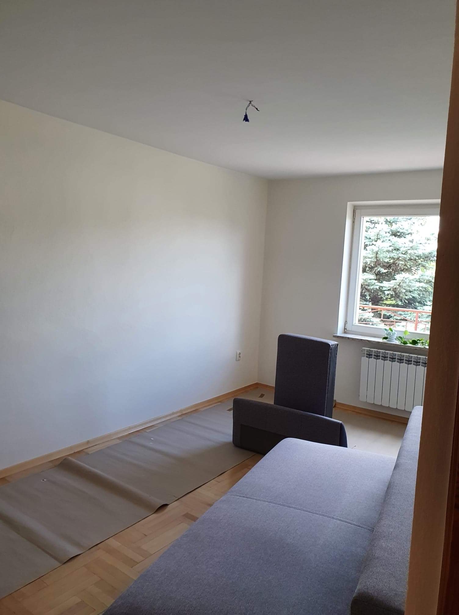 Pokój przygotowany do prac wykończeniowych: podłoga zabezpieczona kartonem, ściany zagruntowane, widoczne kable od lampy sufitowej.