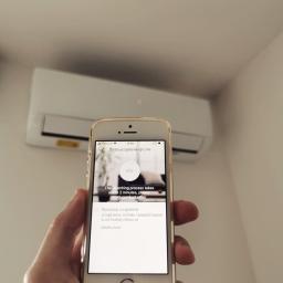 KlimatyzacjaPruszkow - Smartfon w dłoni wyświetla proces dodawania klimatyzatora do aplikacji; w tle widoczny biały klimatyzator ścienny.