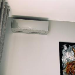 KlimatyzacjaPruszkow - Zamontowany, nowoczesny klimatyzator marki Gree na szarej ścianie w pokoju z zasłoniętym oknem i obrazem sukienki w prawym dolnym rogu.