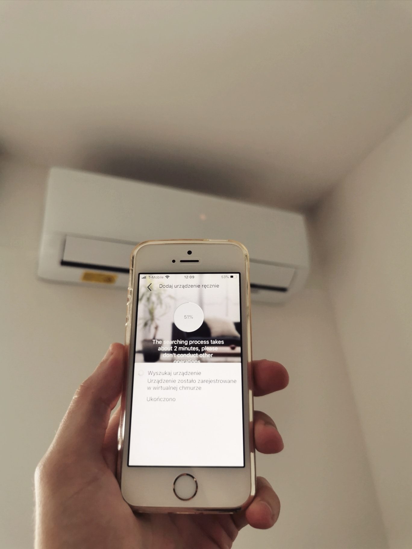 Smartfon w dłoni wyświetla proces dodawania klimatyzatora do aplikacji; w tle widoczny biały klimatyzator ścienny.
