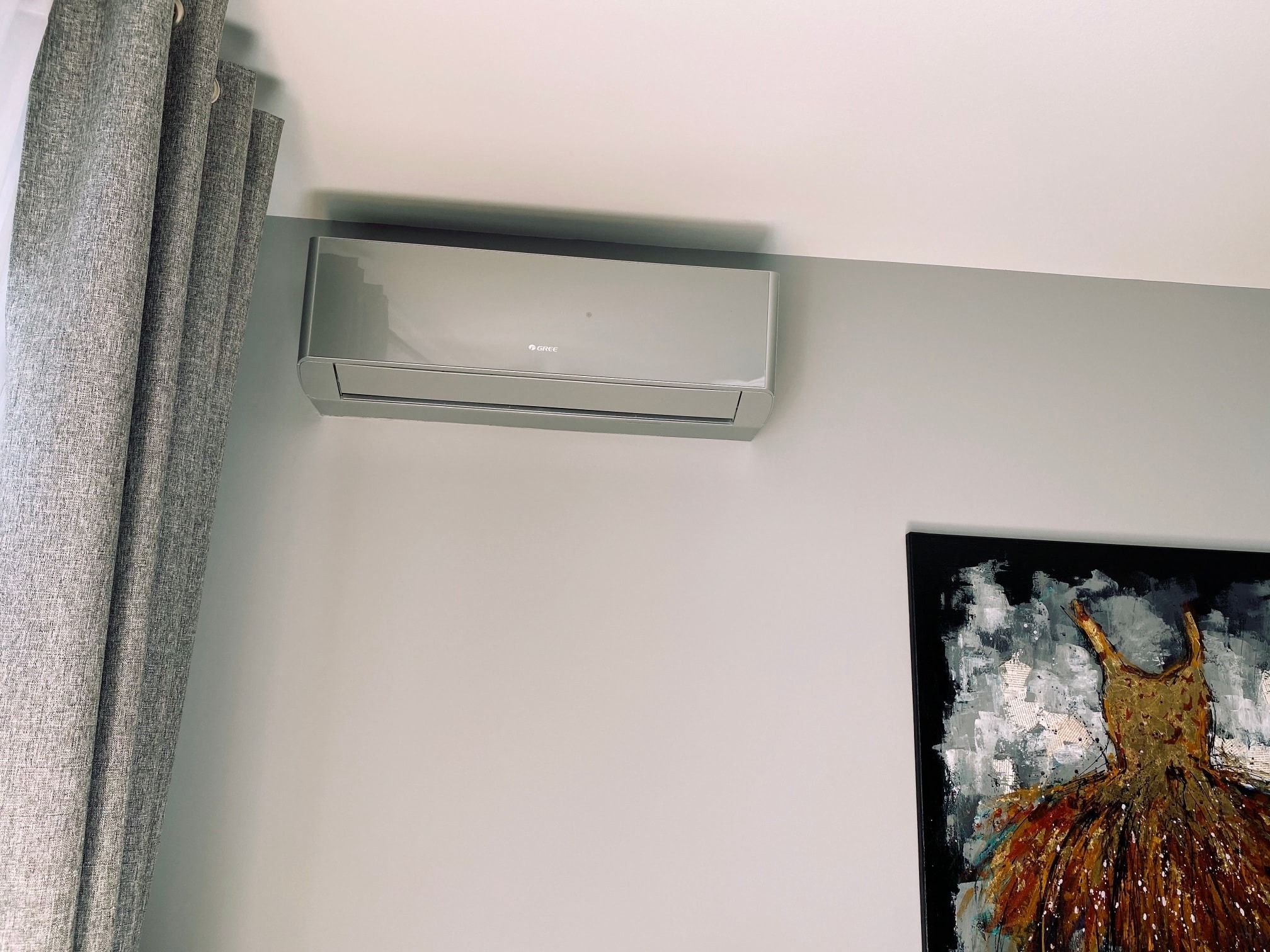 Zamontowany, nowoczesny klimatyzator marki Gree na szarej ścianie w pokoju z zasłoniętym oknem i obrazem sukienki w prawym dolnym rogu.